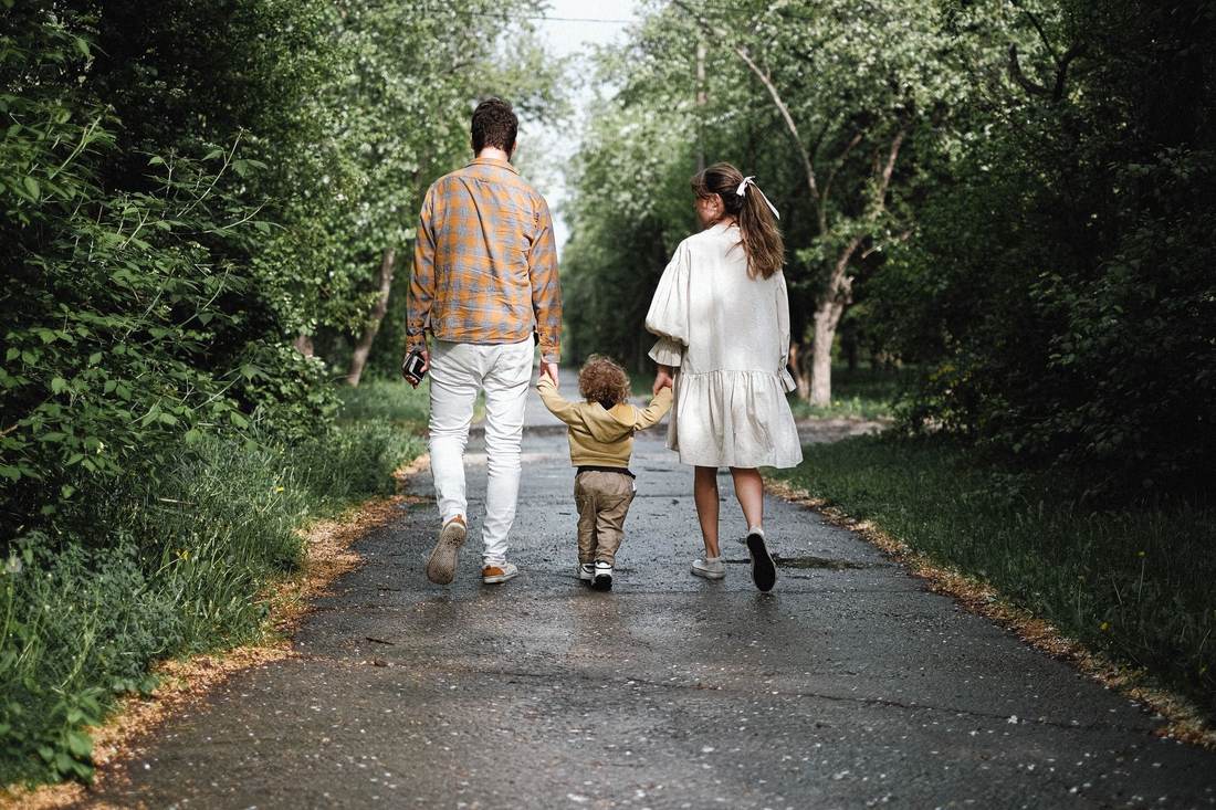 A young family walking hand in hand with their toddler on a tree-lined path