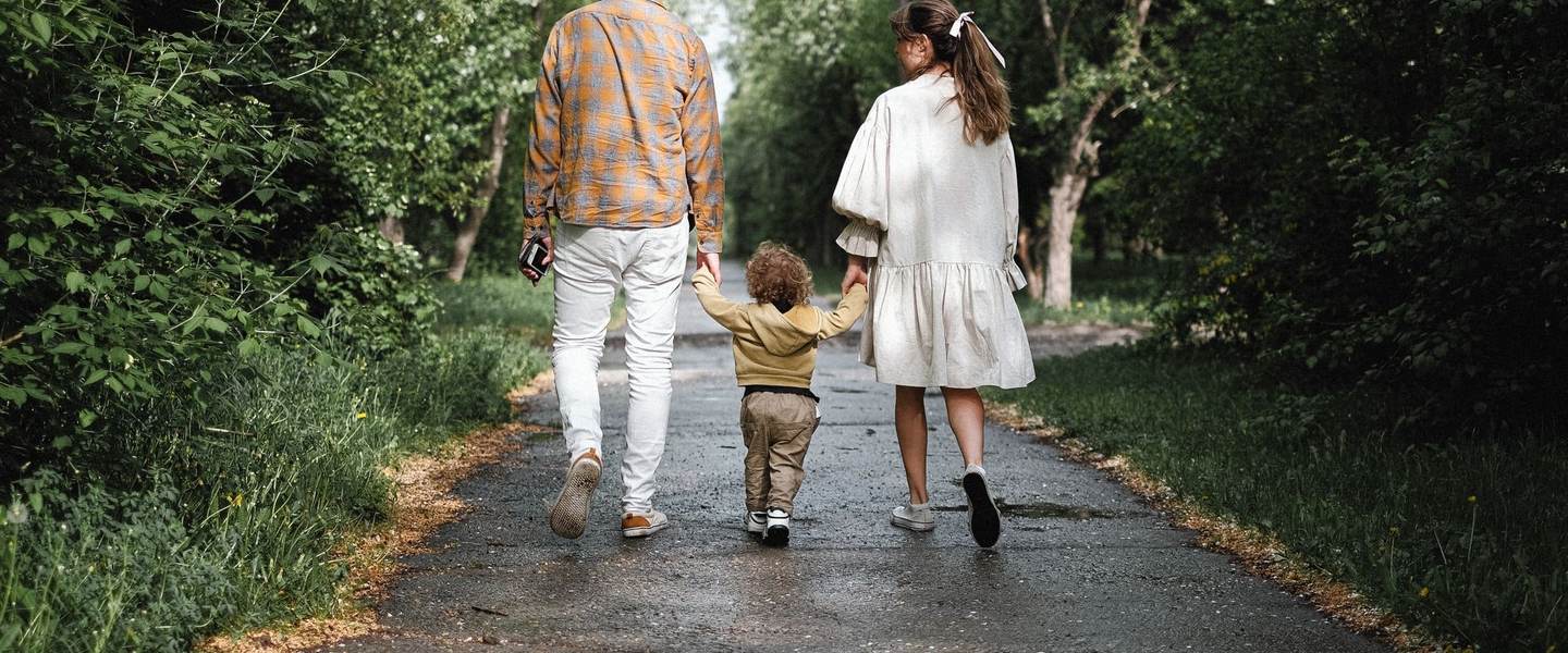 A young family walking hand in hand with their toddler on a tree-lined path